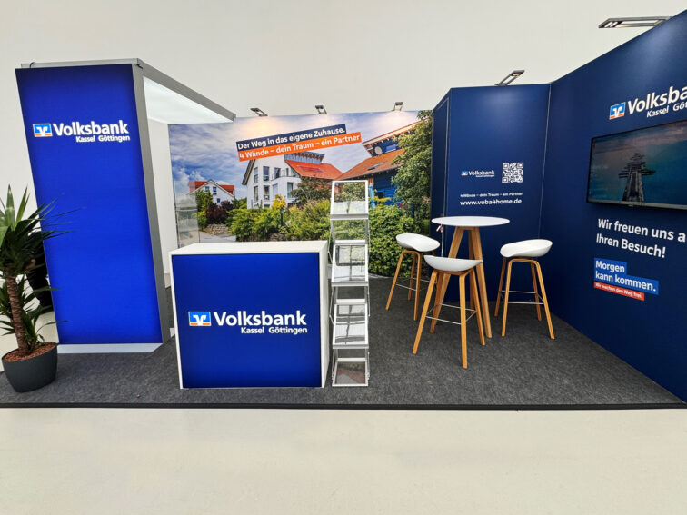 Volksbank Messestand mit Leuchtwänden, Fernseher, Tisch, Stühle und Zeitschriftenständer